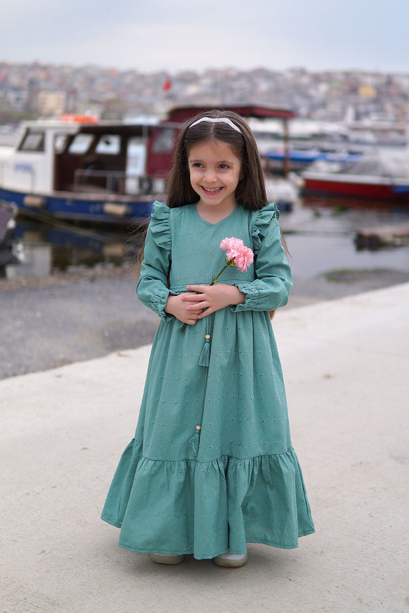 Girl wearing the Dahlia modest maxi dress in teal with ruffled shoulders, holding a pink flower by the harbor