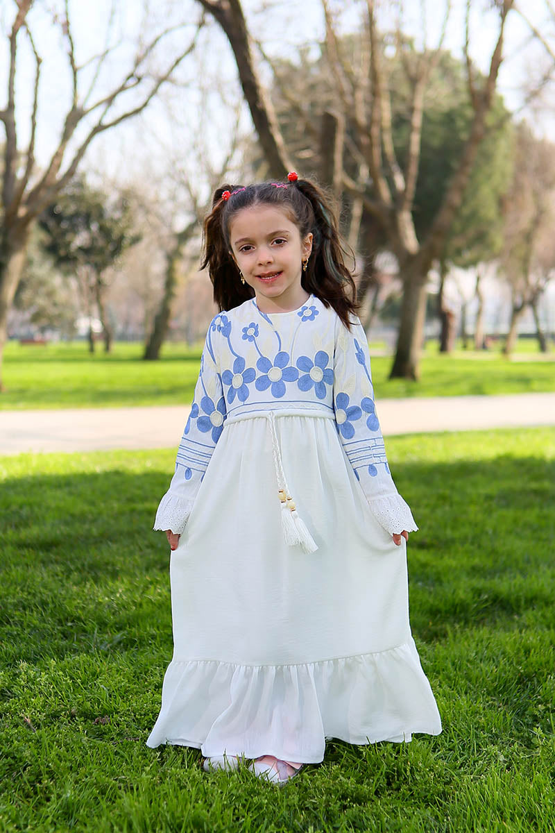 Girl wearing the white linen modest maxi dress with blue daisy embroidered bodice, ruffled cuffs and tassel tie waist